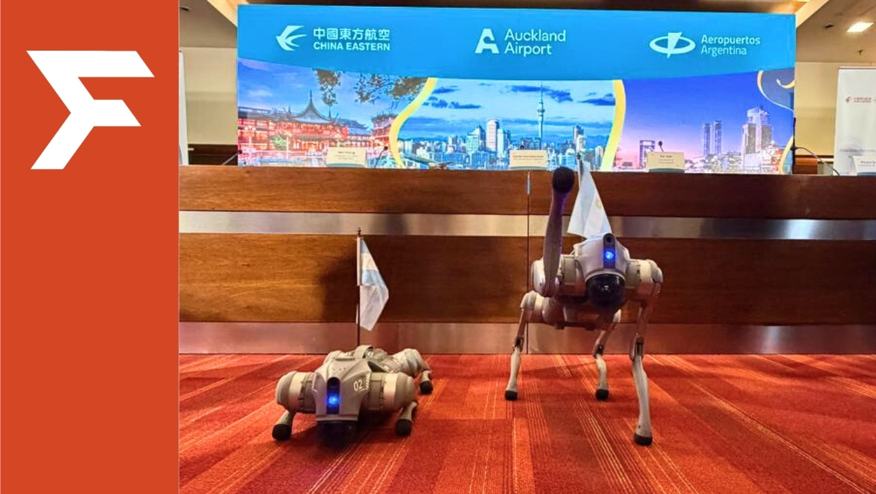Dos robots cuadrúpedos con banderas argentinas en demostración indoor frente a panel de aeropuerto.